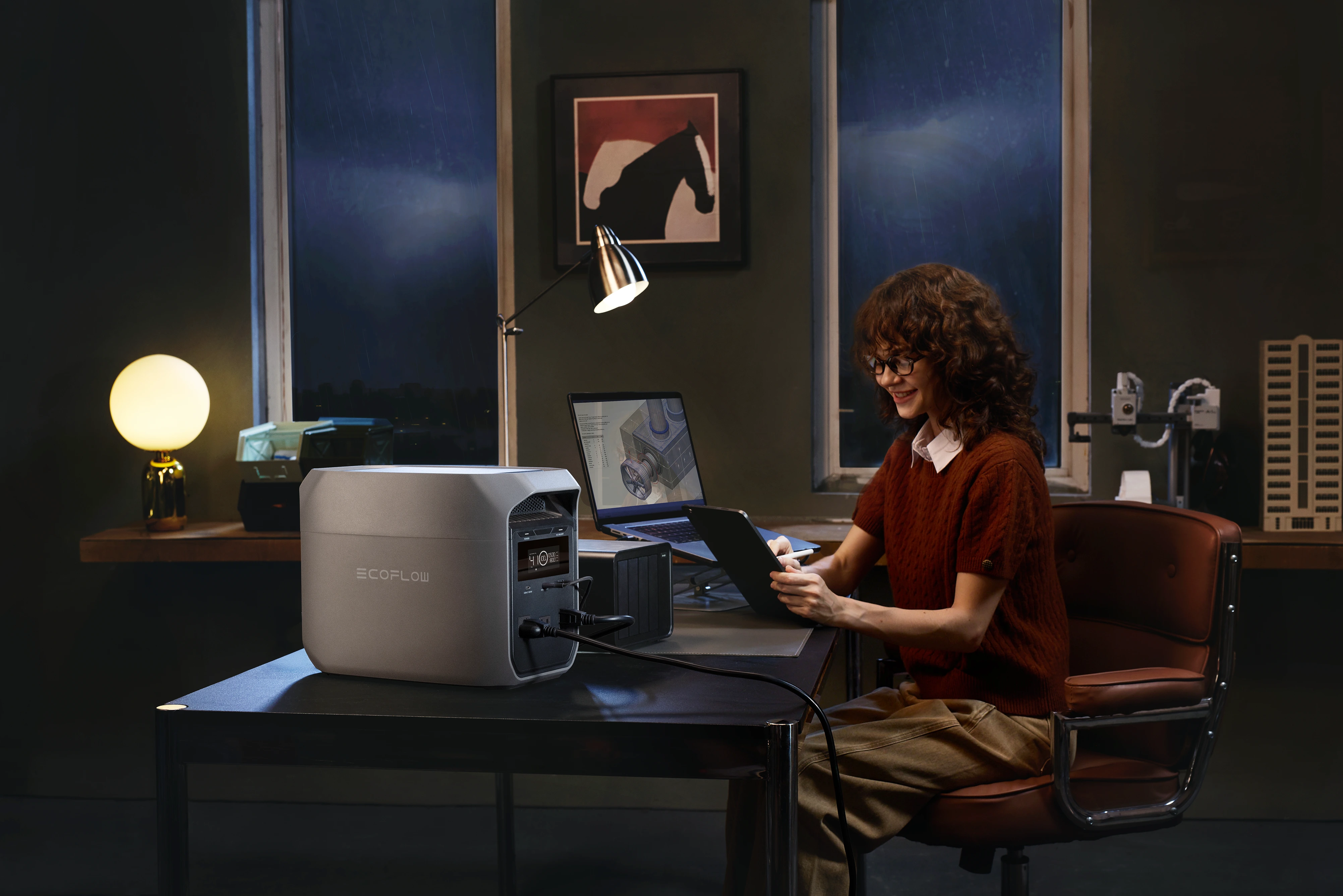 Female designer working on laptop and tablet powered by EcoFlow home power station at night home office, backup battery generator keeps electronics running through storm blackouts